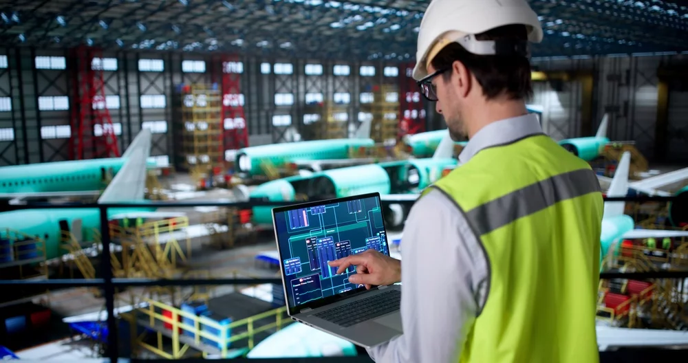 An engineer overlooking an airplane manufacturing center, while typing on a laptop.